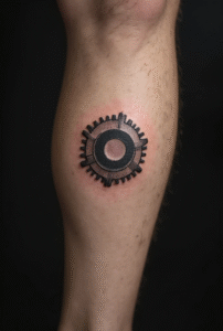 “Small gear icon tattoo on a man’s ankle area, detailed with fine black lines representing progress and hard work.”