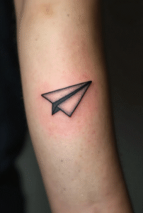 “Small folded paper plane tattoo on a male outer forearm, made with clean minimalist black lines symbolizing freedom and adventure.”